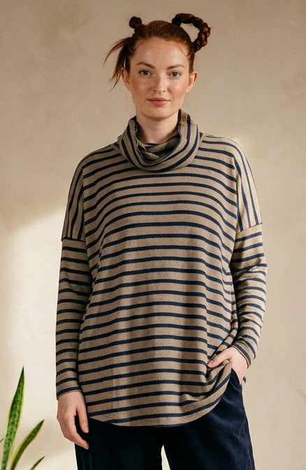 A woman stands in a warm, minimalist room wearing a beige and dark gray striped cowl-neck sweater, dark navy trousers, and black leather shoes. She is posed between potted plants against a textured beige wall.