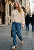 A woman walking on a city street wearing a cream knit sweater, blue jeans, and white sneakers with brown stripes, with historic stone buildings in the background.