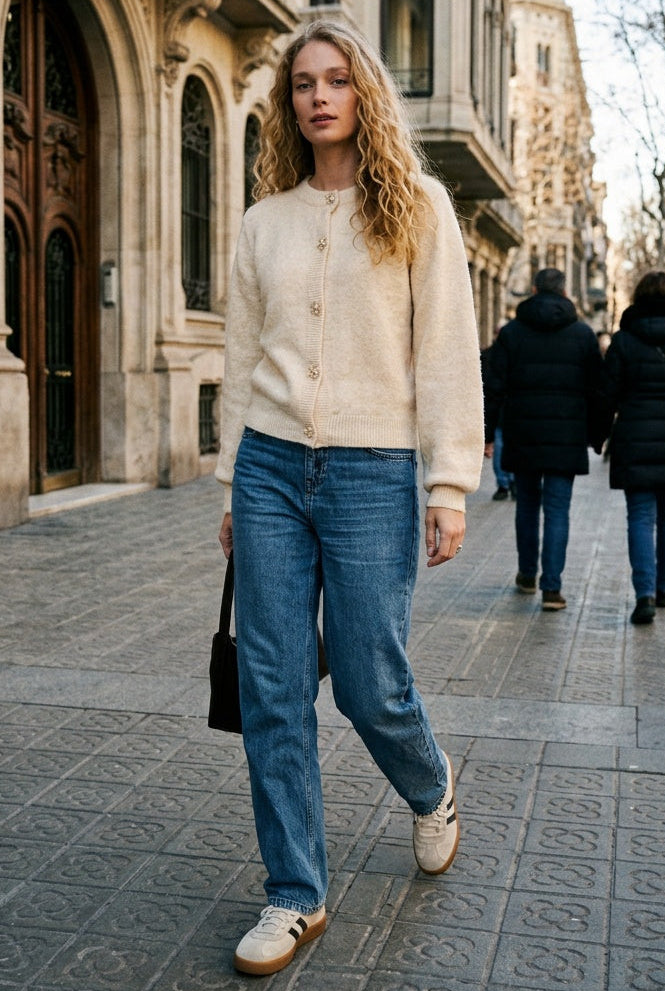A woman walking on a city street wearing a cream knit sweater, blue jeans, and white sneakers with brown stripes, with historic stone buildings in the background.