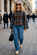 A woman walking on a city sidewalk wearing a brown and beige plaid cropped jacket, blue jeans, white sneakers, and sunglasses, carrying a black handbag.