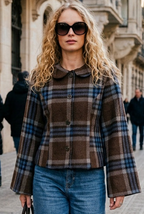 A woman walking on a city sidewalk wearing a brown and beige plaid cropped jacket, blue jeans, white sneakers, and sunglasses, carrying a black handbag.