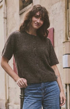 A woman stands on a city sidewalk wearing a dark brown short-sleeve knit top, blue jeans, and beige shoes, posing next to a black metal pole in front of a building with an antique shop window.