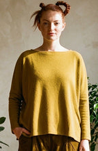 A woman stands in a sunlit room wearing a loose mustard yellow long-sleeve top and matching olive-brown corduroy pants, paired with black shoes. The background features a textured beige wall, a wooden floor, and several potted green plants.