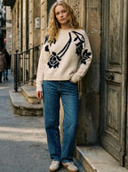 A woman stands on a city sidewalk wearing a cream knit sweater with black floral embroidery, blue jeans, and beige sneakers, leaning against a stone building with a large wooden door.