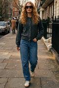 A woman walking down a city street wearing a dark gray knit cardigan, blue jeans, white sneakers, and sunglasses.