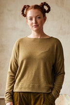 A woman stands in a sunlit room wearing a ribbed long-sleeve top in olive-khaki and matching corduroy pants in a dark brownish-olive. She is styled with black socks and black shoes, with potted plants and a rustic interior background.