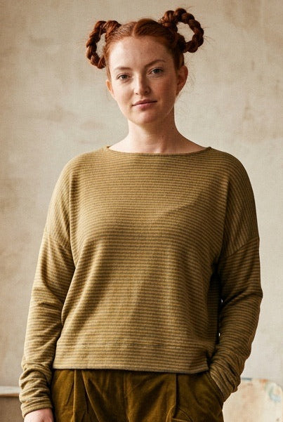 A woman stands in a sunlit room wearing a ribbed long-sleeve top in olive-khaki and matching corduroy pants in a dark brownish-olive. She is styled with black socks and black shoes, with potted plants and a rustic interior background.