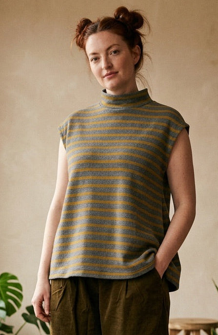 A woman stands in a rustic indoor room wearing a sleeveless mock-neck knit top with muted green and tan horizontal stripes, olive green wide-leg pants, and dark boots. The room has a textured beige wall, wooden floor, and potted plants.