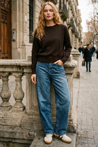 A woman stands on a city sidewalk leaning against a stone balustrade, wearing a dark brown long-sleeve crewneck sweater, wide-leg blue jeans, and white sneakers.