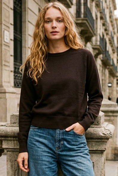 A woman stands on a city sidewalk leaning against a stone balustrade, wearing a dark brown long-sleeve crewneck sweater, wide-leg blue jeans, and white sneakers.
