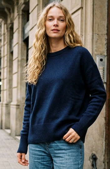 A woman stands on a city sidewalk wearing a navy blue crewneck sweater, loose-fit light-wash blue jeans, and white sneakers with tan soles. The background shows a stone building with a large carved wooden door and other pedestrians in the distance.