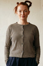 A woman stands in a rustic room wearing a gray and white striped long-sleeve button-up cardigan, dark navy wide-leg pants, and black shoes. The background features a textured off-white wall, a wooden floor, and several potted green plants.