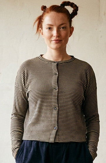 A woman stands in a rustic room wearing a gray and white striped long-sleeve button-up cardigan, dark navy wide-leg pants, and black shoes. The background features a textured off-white wall, a wooden floor, and several potted green plants.