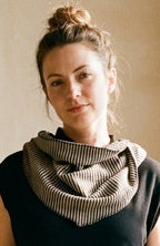 A woman stands in a sunlit room wearing a black sleeveless top, a gray-and-white striped cowl neck scarf, wide-leg gray patterned trousers, and black shoes. Potted plants are arranged against a beige plaster wall.