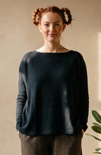 A woman stands in a sunlit room wearing a dark navy long-sleeve top and dark brown patterned trousers, with potted plants on a small wooden table beside her.