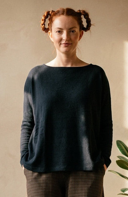A woman stands in a sunlit room wearing a dark navy long-sleeve top and dark brown patterned trousers, with potted plants on a small wooden table beside her.