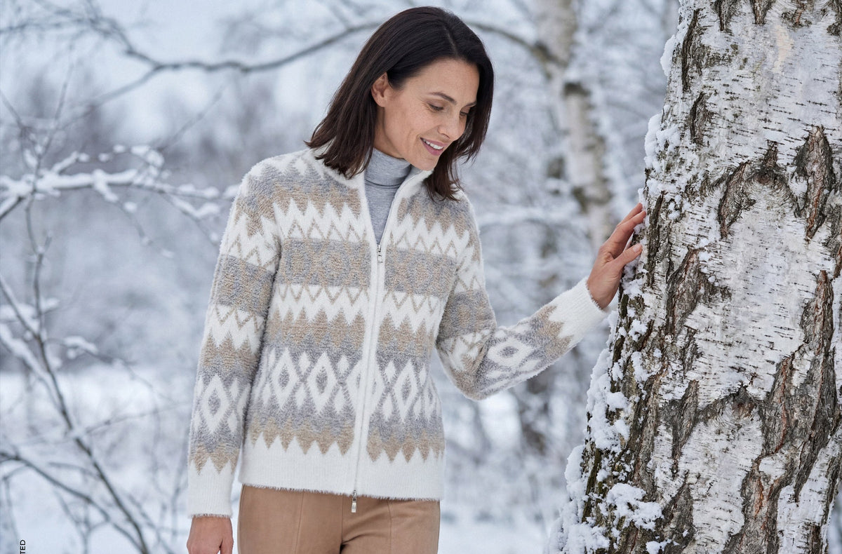 A woman in a snowy outdoor setting wearing a cream and beige geometric patterned zip-up cardigan sweater, standing next to a birch tree.