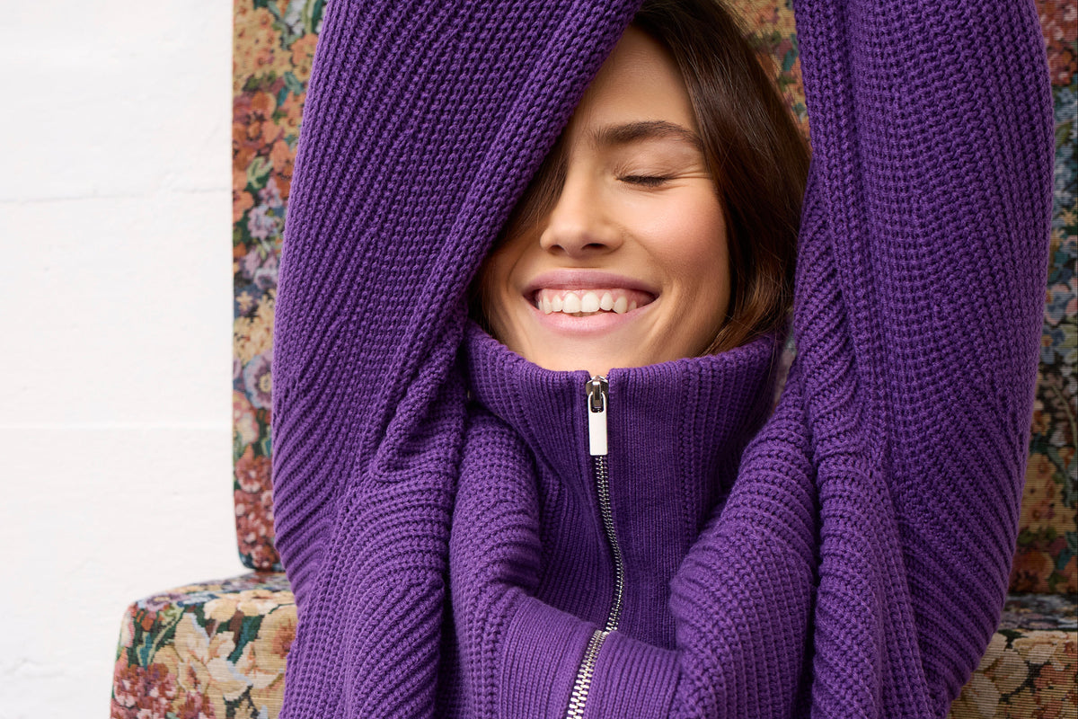 A woman smiling with her eyes closed, wrapped in a cozy purple ribbed knit zip-up sweater draped over her head and shoulders, seated on a floral armchair.