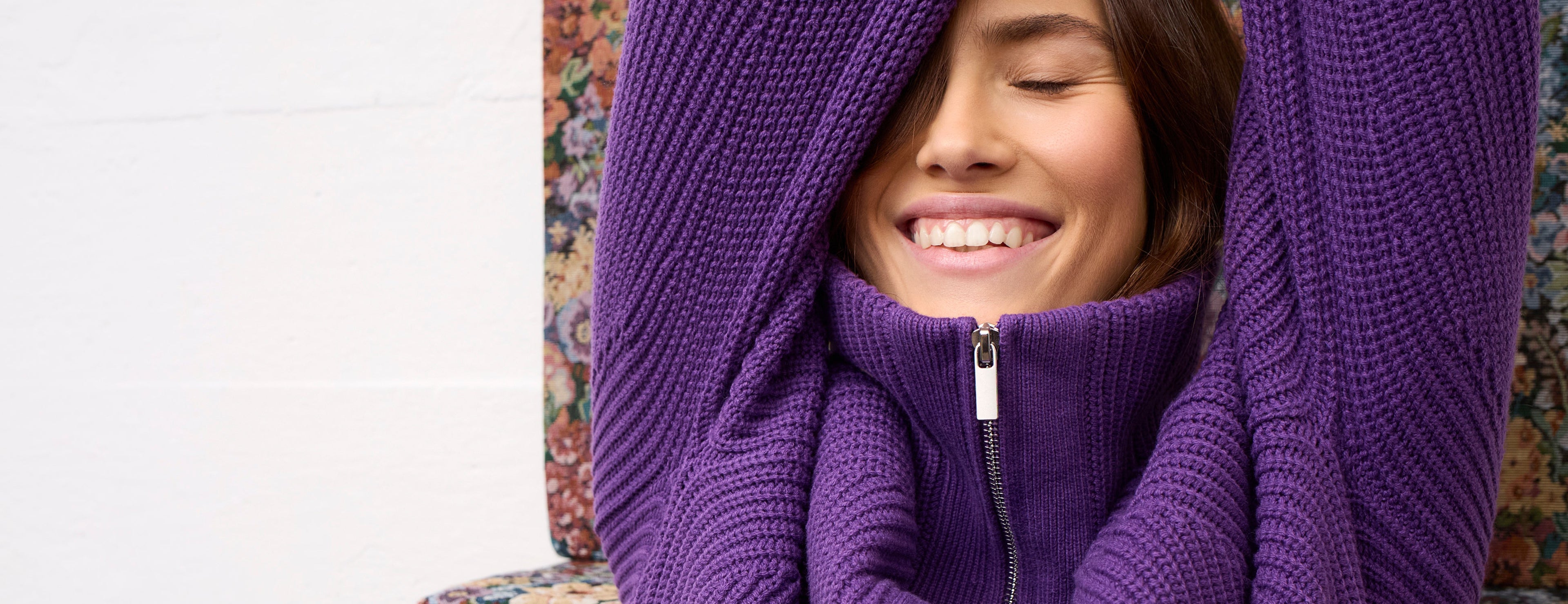 A woman smiling with her eyes closed, wrapped in a cozy purple ribbed knit zip-up sweater draped over her head and shoulders, seated on a floral armchair.
